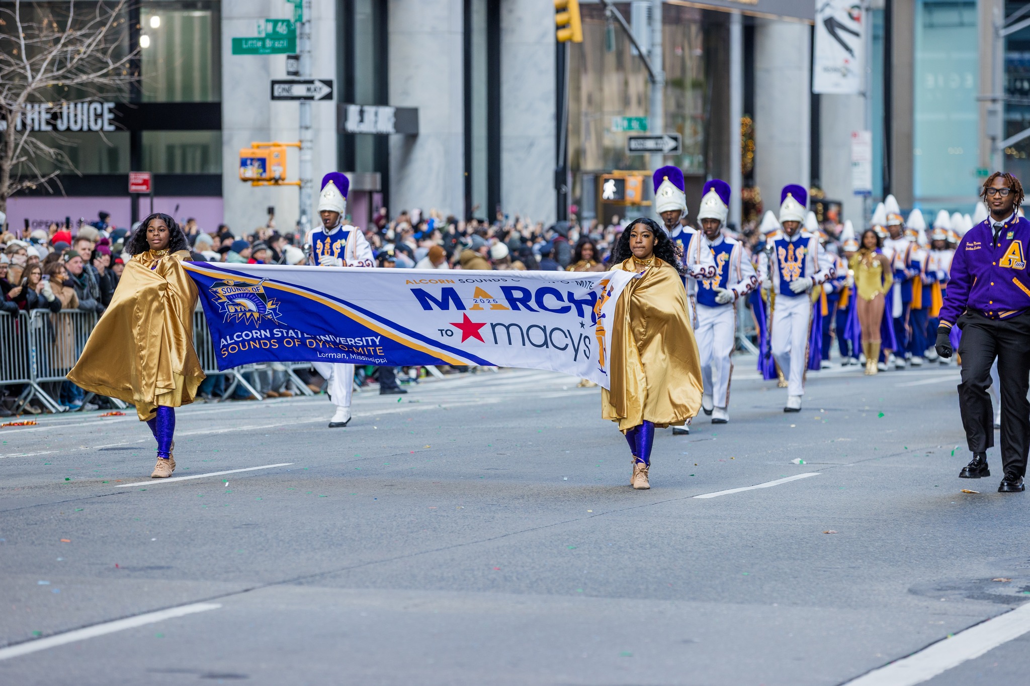 Two women in gold capes carry an “Alcorn Sounds of Dyn-O-Mite March to Macy’s” banner while the Alcorn State University marching band follows behind them on a New York City street during the 2025 Macy’s Thanksgiving Day Parade.